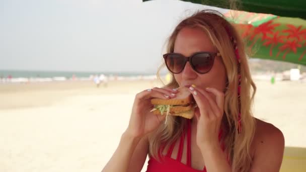 Una joven en un café en la playa se come un sándwich. — Vídeo de stock