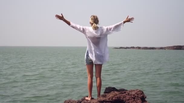 Női turista áll egy sziklán a tenger felett, és széttárja a karját az oldalán — Stock videók