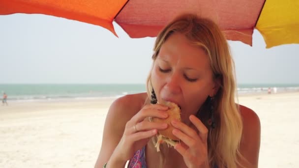 Young woman eating a hamburger in a cafe on the beach. Improper nutrition, fast food — Stock Video