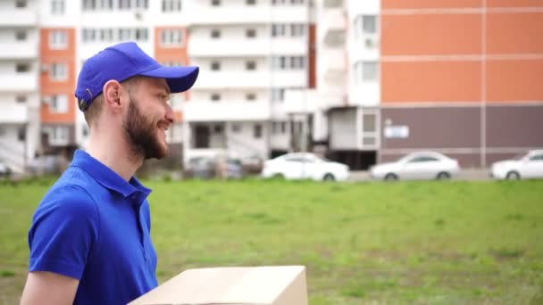 Jovem mensageiro positivo carrega uma caixa de papelão. Carteiro, empregado do serviço de entrega — Vídeo de Stock