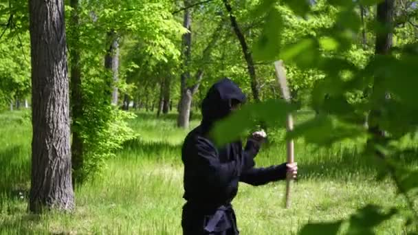 Japonský ninja trénuje s dřevěnou holí s obuškem. Ninja v obleku v lese — Stock video
