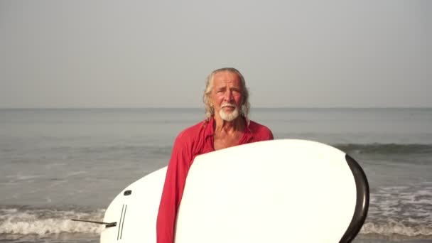 Een volwassen mannelijke gepensioneerde, een oude man met een surfplank komt uit de zee — Stockvideo