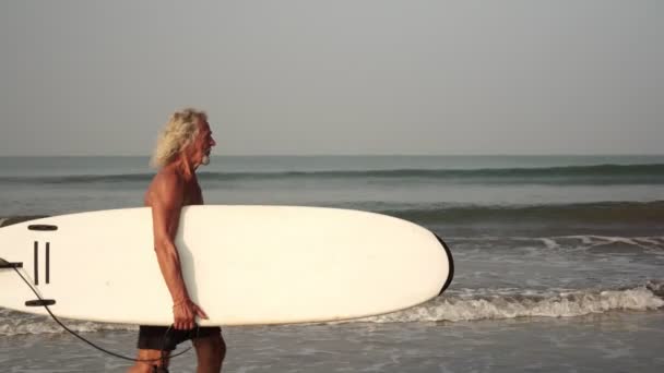 Volwassen mannelijke surfer. Grijsharige oude man opa op het strand met een surfplank — Stockvideo