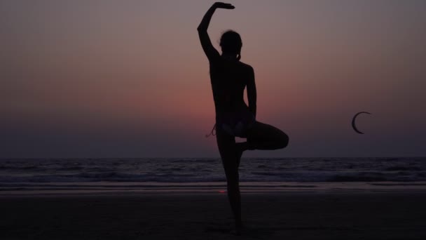 女性は一本足で立ち、ビーチで日没時にヨガを練習します — ストック動画