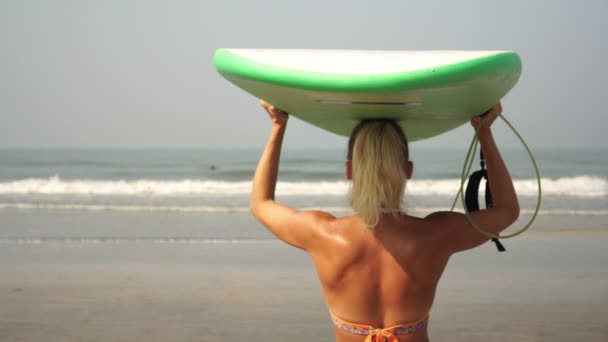 Uma jovem surfista vai para o mar com uma prancha de surf. Uma linda garota está surfando — Vídeo de Stock