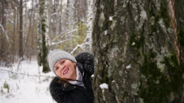 Девушка играет, бросая снежки из-за дерева в зимнем парке — стоковое видео
