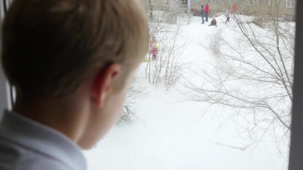 Um adolescente olha pela janela para a queda de neve e brincando de crianças — Vídeo de Stock
