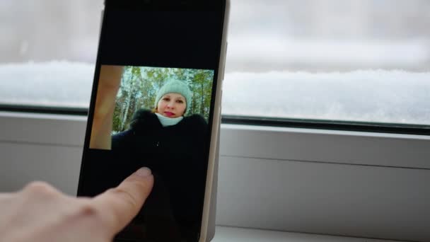 Chica bebiendo té cerca de la ventana y viendo sus fotos en el teléfono inteligente — Vídeos de Stock