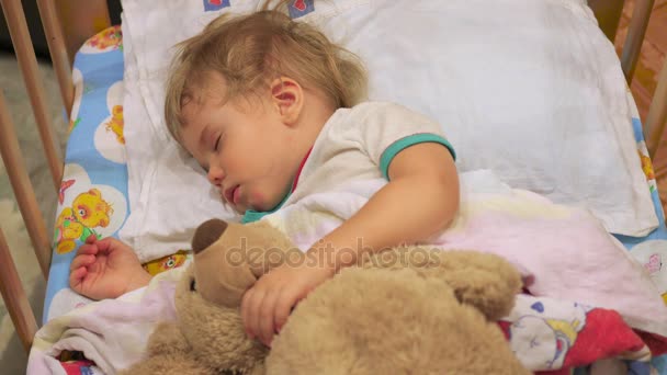The sweet baby sleeps in a cot with a teddy bear. — Stock Video