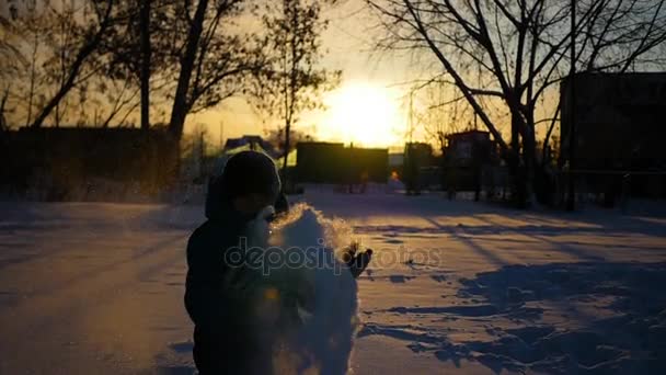 G niño lanzando nieve sobre sí mismo y disfruta de ella en el parque de invierno al atardecer — Vídeo de stock