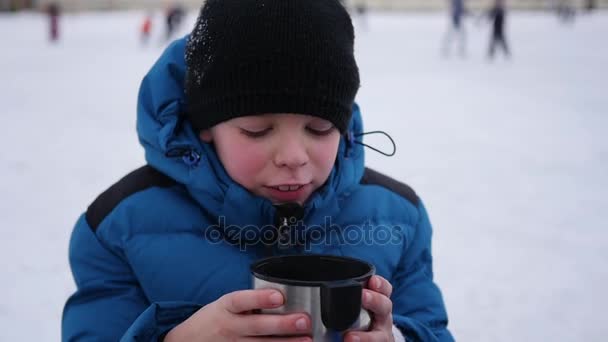 The kid is drinking hot tea from a mug in the winter outdoor. ice rink. — Stock Video