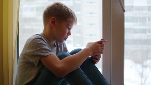 Das Kind sitzt auf der Fensterbank und schaut auf das Smartphone — Stockvideo