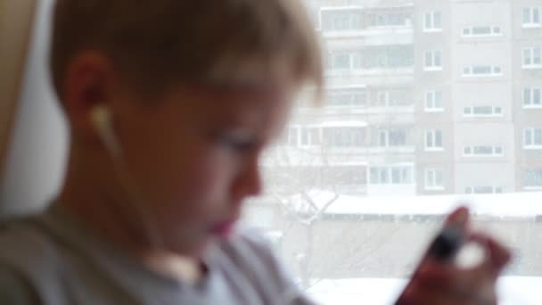 L'enfant s'assoit sur le rebord de la fenêtre et écoute de la musique sur smartphone dans un casque — Video