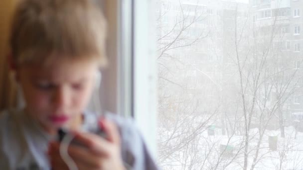 L'enfant s'assoit sur le rebord de la fenêtre et écoute de la musique sur smartphone dans un casque — Video