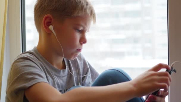 Il bambino si siede sul davanzale della finestra e ascolta musica allo smartphone in primo piano con le cuffie — Video Stock