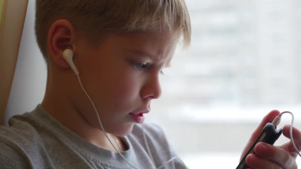 Ребенок сидит на подоконнике и слушает музыку на смартфоне в наушниках крупным планом — стоковое видео
