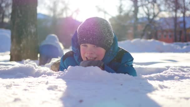 Fröhliches Kind, das an einem sonnigen Tag im Winterpark Spaß hat.Liegend im Schnee — Stockvideo