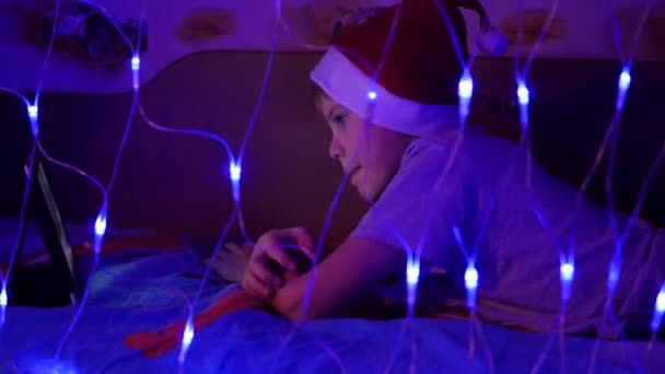 Dítě vypadá na tabletu, ležící na posteli v klobouk santa. V popředí světla girlandy — Stock video