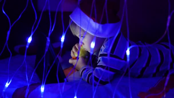 The child looks to the tablet lying on bed in hat santa.On the foreground, lights garlands — Stock Video