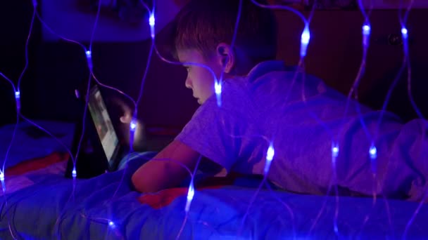 The child looks to the tablet lying on bed.On the foreground, lights garlands — Stock Video