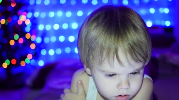 El niño mira a la tableta acostada en la cama. En el fondo, luces y guirnaldas de abeto navideño — Vídeos de Stock