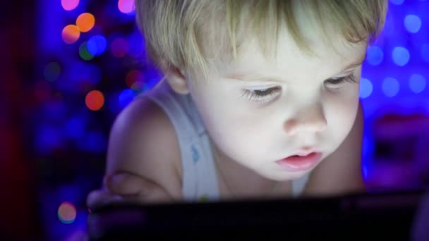 Dítě vypadá na tabletu, ležící na posteli. V pozadí, světla a girlandy vánoční jedle — Stock video
