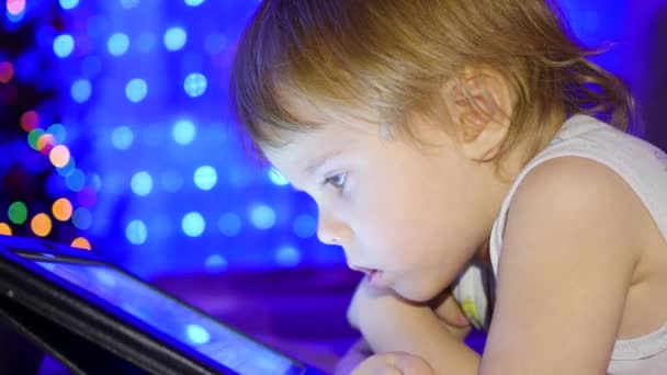 Das Kind blickt auf das Tablet, das auf dem Bett liegt. im Hintergrund Lichter und Girlanden aus Weihnachtstannen — Stockvideo