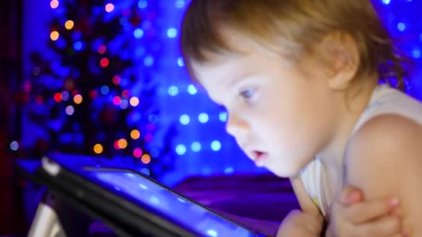 Het kind kijkt naar de tablet liggend op bed. In de achtergrond, de lichten en de slingers van Kerstmis spar — Stockvideo
