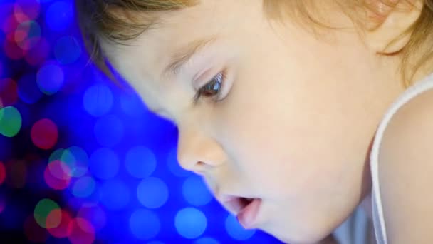 Het kind kijkt naar de tablet liggend op bed. In de achtergrond, de lichten en de slingers van Kerstmis spar. Closeup — Stockvideo