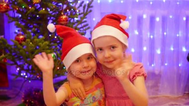 Gülümseyen ve Noel Baba şapkası kamerada, sallayarak iki sevimli kız. Arka plan, ışıklar ve Noel köknar çelenk