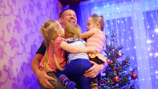 Feliz padre sostiene en las manos de los niños, sonriendo y riendo en una noche de Navidad. En el fondo, luces y guirnaldas de abeto navideño — Vídeo de stock