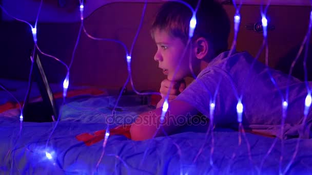 L'enfant regarde la tablette couchée sur le lit.Au premier plan, des guirlandes de lumières — Video