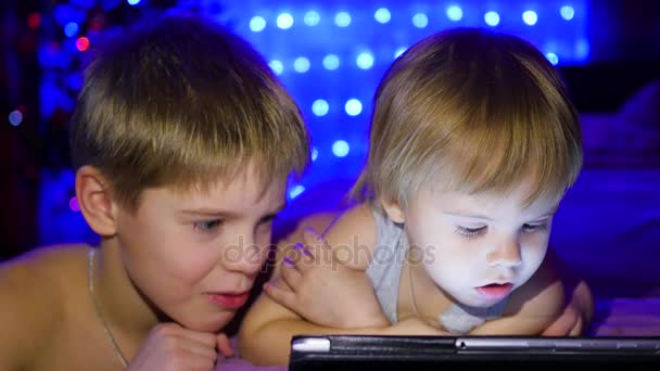 Los niños miran el plato acostado en la cama. En el fondo, luces y guirnalda abeto de Navidad — Vídeo de stock