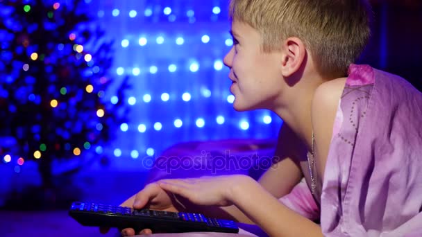 El niño mira al televisor acostado en la cama. En el fondo, luces y guirnaldas de abeto navideño — Vídeos de Stock