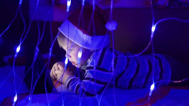 Il bambino guarda la tavoletta sdraiato sul letto in cappello santa.On in primo piano, ghirlande luci — Video Stock