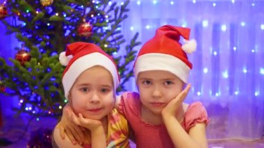Gülümseyen ve Noel Baba şapkası kamerada, sallayarak iki sevimli kız. Arka plan, ışıklar ve Noel köknar çelenk