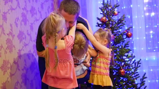Szczęśliwy ojciec trzyma w ręce dzieci, uśmiecha się i śmieje się w wigilijny wieczór. W tle, światła i Girlandy choinkowe Christmas — Wideo stockowe