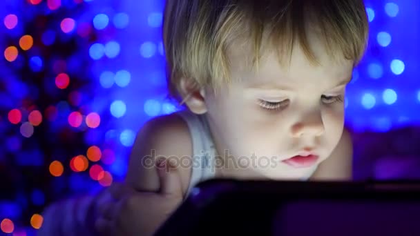 Das Kind blickt auf das Tablet, das auf dem Bett liegt. im Hintergrund Lichter und Girlanden aus Weihnachtstannen — Stockvideo