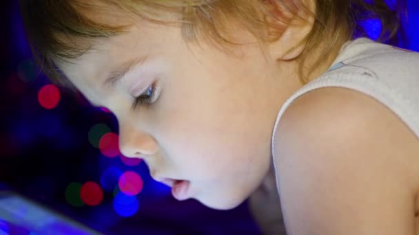 El niño mira a la tableta acostada en la cama. En el fondo, luces bokeh y guirnaldas de abeto de Navidad — Vídeos de Stock