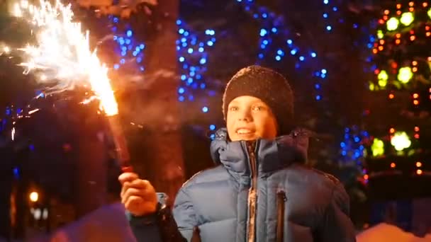 El niño sostiene los fuegos artificiales al aire libre en el invierno. En cámara lenta. En el fondo, luces y guirnaldas de abeto navideño — Vídeos de Stock