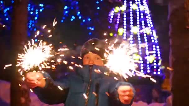 Дитина тримає блискавки на відкритому повітрі взимку. Повільність. На задньому плані, вогні та гірлянди різдвяної ялинки — стокове відео