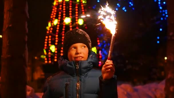 Ребенок держит фейерверк на улице зимой. Медленно. На заднем плане гирлянды и гирлянды рождественской ели — стоковое видео