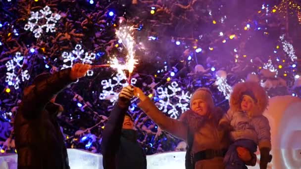 A família mantém os fogos de artifício ao ar livre no inverno. No fundo, luzes e guirlandas de abeto de Natal — Vídeo de Stock