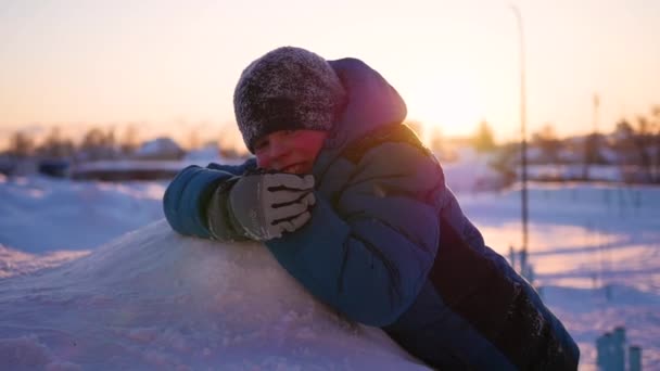 Das Kind liegt auf Schnee und schaut in den Sonnenuntergang. Winterpark — Stockvideo