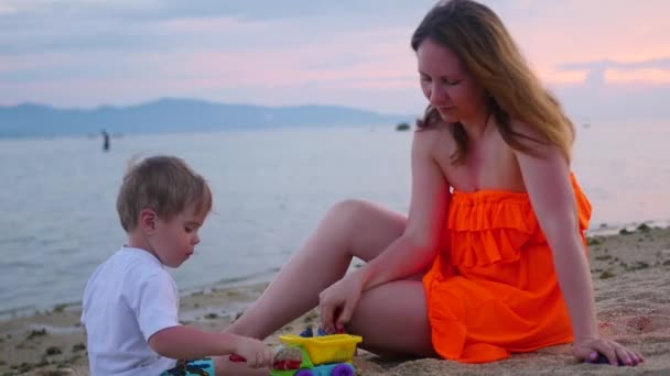 Chica con un niño juega con arena en la playa al atardecer — Vídeos de Stock