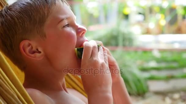 El niño está comiendo sandía jugosa dulce en un día caliente del verano que miente en una hamaca — Vídeos de Stock