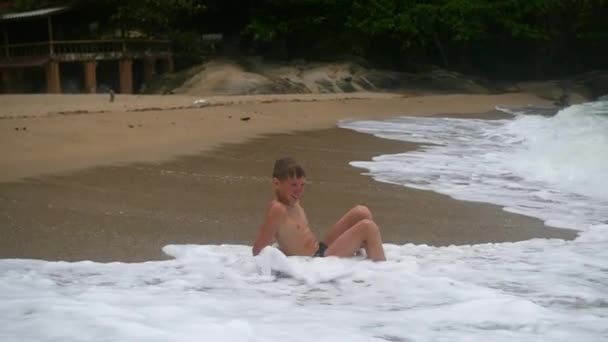 El niño se encuentra en la playa y está bañado en grandes olas marinas — Vídeos de Stock