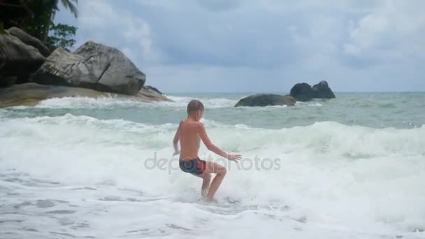 El niño se encuentra en la playa y está bañado en grandes olas marinas — Vídeos de Stock