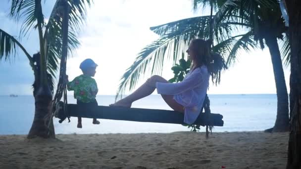 A girl and a child swinging on a rope swing on the beach — Stock Video