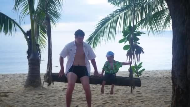 Un chico y un niño balanceándose en un columpio de cuerda en la playa — Vídeos de Stock